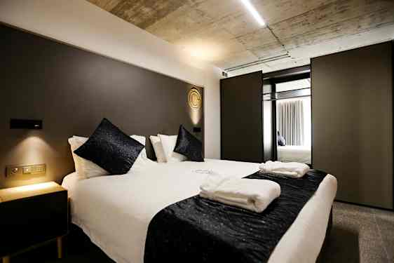 Sovrum med en modern dubbelsäng, vita sängkläder och mörka dekorativa kuddar. En nattduksbord med mjuk belysning står bredvid. (AI-genererad text)