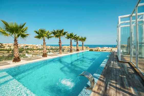 En utomhuspool med turkost vatten, omgiven av palmer och med utsikt över en stad och havet. (AI-genererad text)