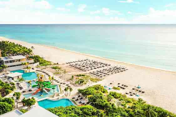Bild av en sandstrand vid havet med blått vatten. På stranden finns solstolar och parasoller. I förgrunden syns en pool med vattenrutschkanor samt omgivande gröna växter. (AI-genererad text)