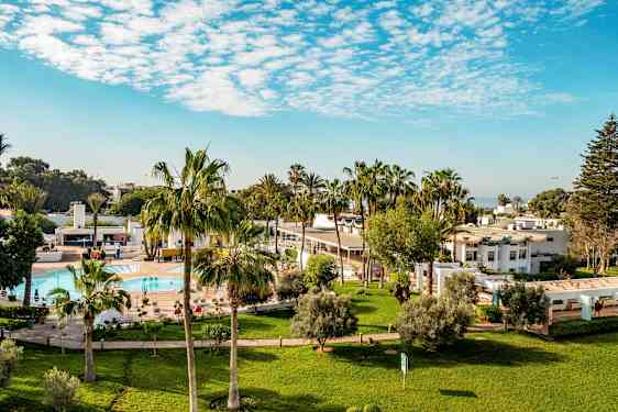 Hotellområde med gräsmattor, palmer, pool och vita byggnader. (AI-genererad text)