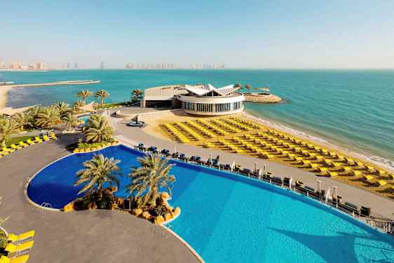 En bild av ett modernt hotellkomplex vid havet. I förgrunden syns en stor pool och solstolar på en strand. (AI-genererad text)