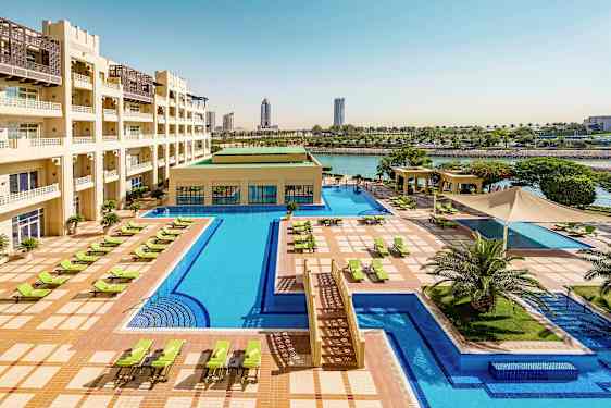 Ett hotellområde med en stor pool, solstolar och gröna uterum. Byggnaden har flera våningar och ligger vid vattnet. (AI-genererad text)