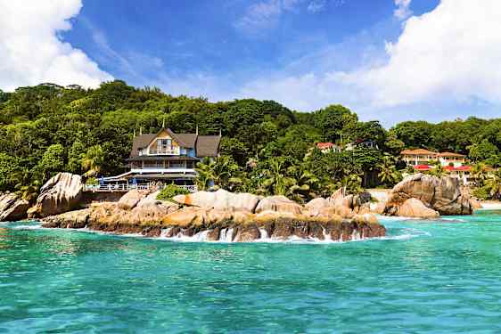 Strand med klippor framför en byggnad omgiven av grönska. (AI-genererad text)