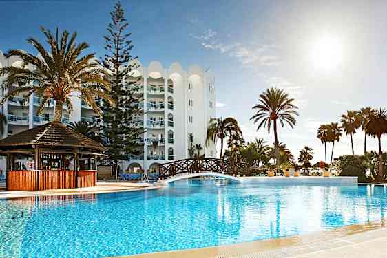 En utomhuspool vid ett hotell, med palmer och en träbro över poolen. (AI-genererad text)