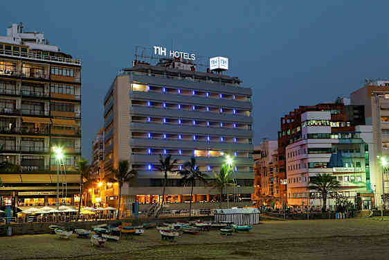 Bild av ett hotell vid havet på kvällen, med ljusskyltar och omgivande byggnader. (AI-genererad text)