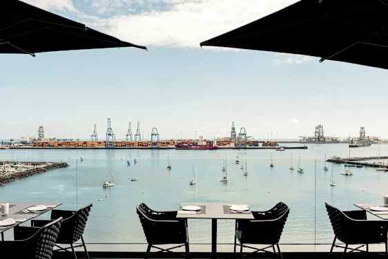 Utsikt över en hamn med kranar och förtöjda båtar. Förgrunden visar bord och stolar från ett café. (AI-genererad text)