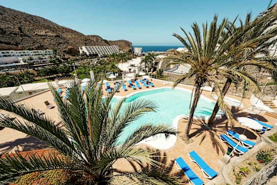 Vy över en hotellpool omgiven av solstolar och palmer med berg och hav i bakgrunden. (AI-genererad text)