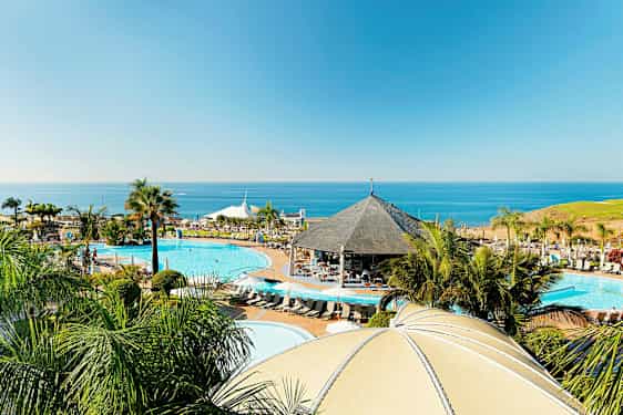 Ett resortområde vid kusten med pooler, en paviljong och palmer, med havet i bakgrunden. (AI-genererad text)