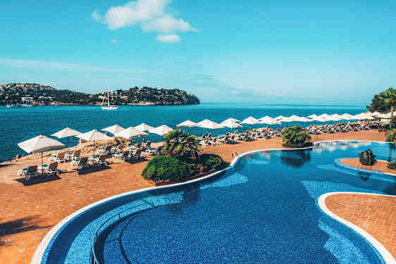 En pool med blått kakel ligger intill en terrass med solsängar och parasoller, med utsikt över en havsvik. (AI-genererad text)