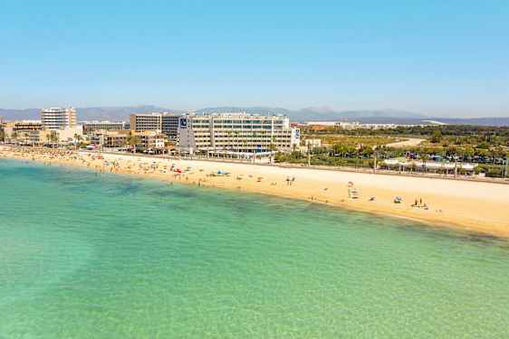 Sandstrand med badande personer och klara turkosa vatten. Hotell och byggnader i bakgrunden. (AI-genererad text)