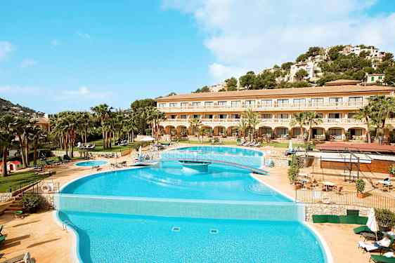 Bilden visar en hotell med en stor pool framför och omgivande terrass med solstolar. Bakgrunden har en grönskande kulle och klara blå himmel. (AI-genererad text)