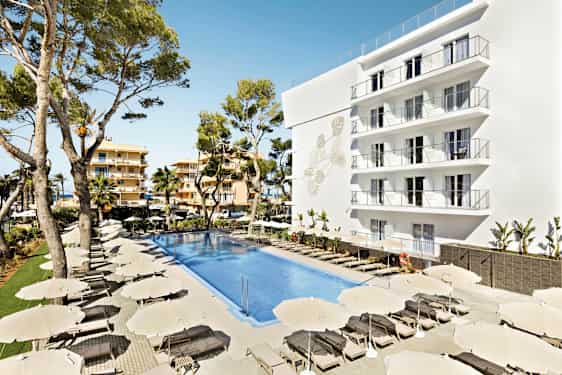 Bild av ett hotellområde med en stor pool omgiven av solstolar och parasoller. En vit byggnad med flera balkonger samt träd i bakgrunden. (AI-genererad text)