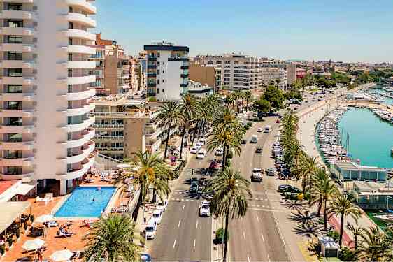 Bild som visar en gata med palmträd, en pool vid ett byggnadskomplex och en hamn med båtar. (AI-genererad text)