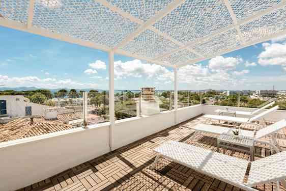 En takterrass med solstolar och ett bord. Terrassen har ett perforerat takskydd och glasräcke med utsikt över stad och hav. (AI-genererad text)