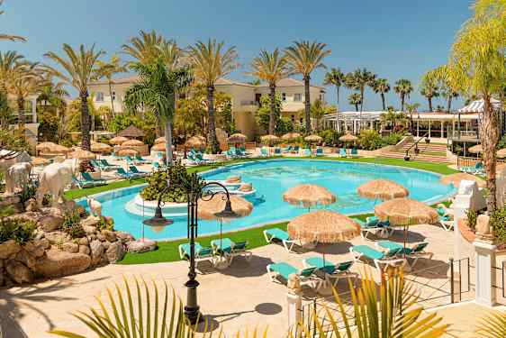 En bild av en lyxig semesteranläggning med en pool, omgiven av solstolar och parasoll, omgivna av palmer och byggnader. (AI-genererad text)