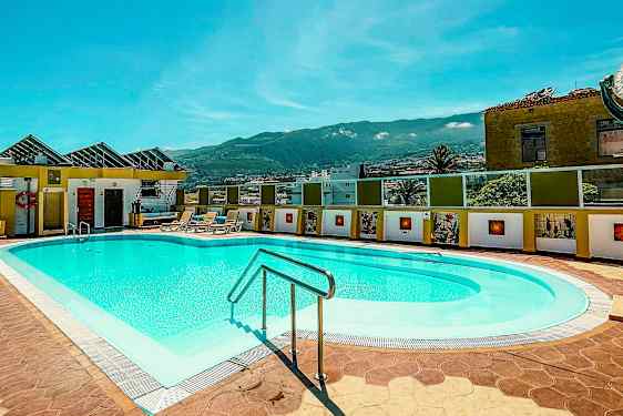 Utomhuspool med solstolar i en solig miljö, omgiven av byggnader och med berg i bakgrunden. (AI-genererad text)