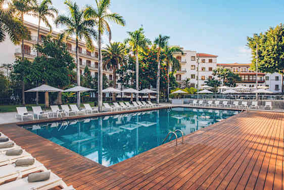 En utomhuspool omgiven av solstolar, parasoll och grönskande träd, med en byggnad i bakgrunden. (AI-genererad text)