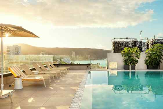 Utsikt över en pool med solstolar och parasoll, i bakgrunden syns en stad och hav. (AI-genererad text)