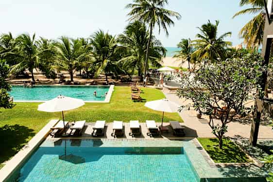 Bilden visar en hotellets poolområde med solstolar och parasoll, omgivna av tropiska palmer, med utsikt mot stranden och havet. (AI-genererad text)