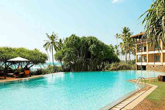 En klarblå pool med växtlighet och palmer i bakgrunden samt en byggnad till höger. (AI-genererad text)