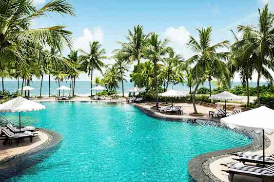 Bild av en pool omgiven av palmer och solsängar med parasoll vid havet. (AI-genererad text)