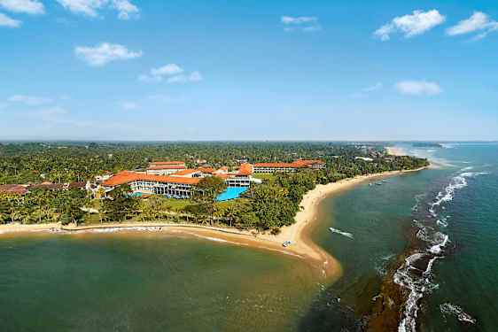 Ett flygfoto av en kustlinje med sandstrand, hav och byggnader omgivna av grönska. (AI-genererad text)