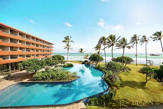 En hotellbyggnad vid stranden med en stor pool, palmer och hav i bakgrunden. (AI-genererad text)