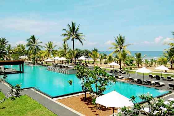 En pool utomhus omgiven av solstolar, parasoller, palmer och med utsikt över havet. (AI-genererad text)