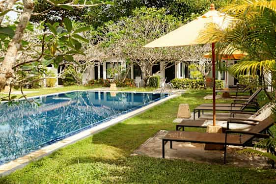 En pool med grönområden och solstolar bredvid, samt en rad vita byggnader i bakgrunden. (AI-genererad text)