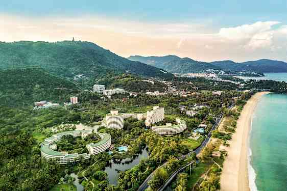 Flygfoto av ett kustnära område med hotellbyggnader, omgivande grönska och en strand. (AI-genererad text)