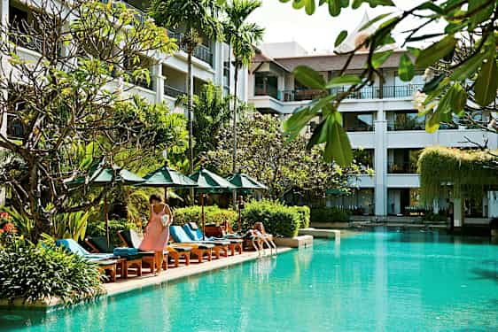 Bild av en utomhuspool omgiven av solstolar, parasoller och växter, med en byggnad i bakgrunden med balkonger. (AI-genererad text)