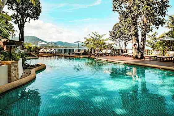 Utomhuspool med klart vatten omgiven av solstolar, parasoller och grönska. Utsikten visar berg och hav i bakgrunden. (AI-genererad text)