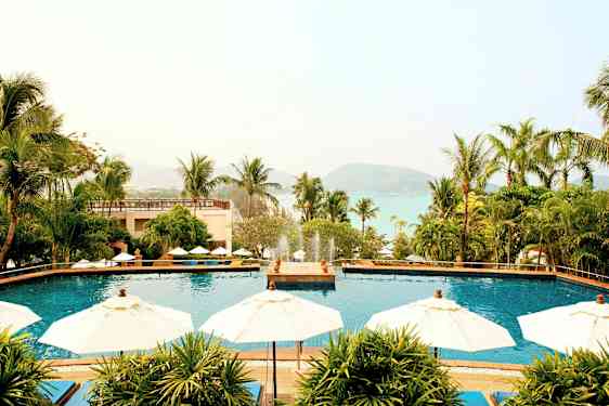 Bild av en semesteranläggning med en utomhuspool omgiven av parasoller och palmer, med utsikt mot havet och berg i bakgrunden. (AI-genererad text)