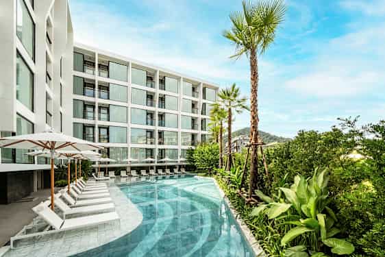 Bild av en modern vit byggnad med stora fönster. Framför byggnaden finns en pool omgiven av solstolar och parasoller, samt palmer och grönska. (AI-genererad text)