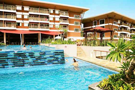 Ett hotell med två våningar syns bakom en utomhuspool där människor simmar. Gröna växter och terrassområden omger poolområdet. (AI-genererad text)