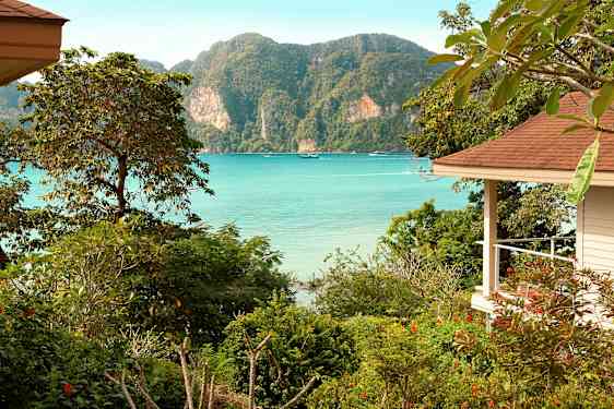 En vy över en havsvik med klarblått vatten, omgiven av grönskande växtlighet och berg, med en del av en byggnad till höger. (AI-genererad text)