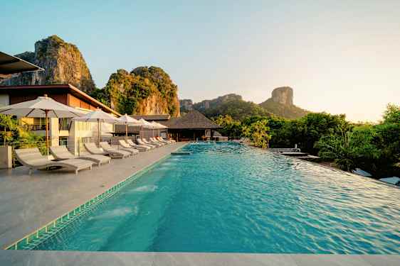 En pool med solstolar och parasoller vid en resort belägen i en naturskön miljö med berg i bakgrunden. (AI-genererad text)