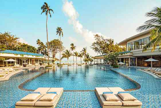 En stor utomhuspool omgiven av solstolar, palmer och byggnader, med utsikt mot havet. (AI-genererad text)