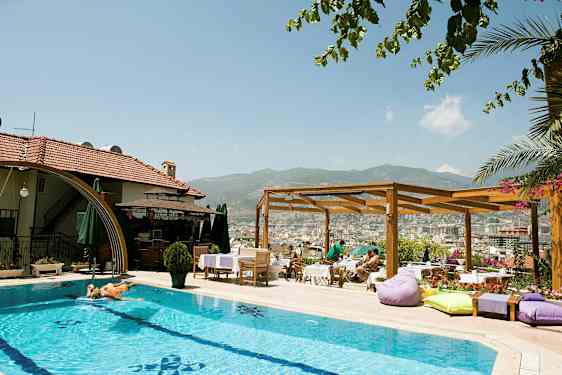 En utomhuspool vid ett hotell med solstolar och parasoller. En träpergola och utsikt över en stad och berg i bakgrunden. (AI-genererad text)