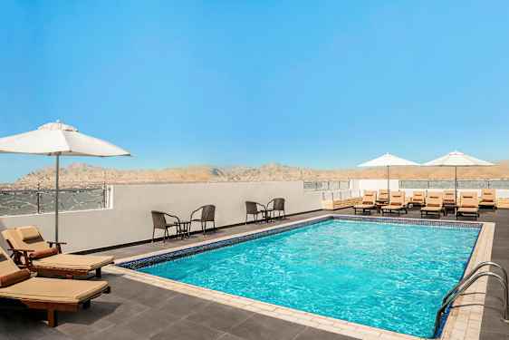 En utomhuspool omgiven av solstolar med parasoller och sittgrupper, med en utsikt över berg och en klarblå himmel. (AI-genererad text)