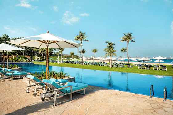 En pool med solstolar och parasoller i en utomhusmiljö, omgiven av palmer och gräsmatta. (AI-genererad text)