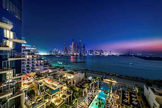 Vy över en modern stadsmiljö vid skymning med skyskrapor, hav och en byggnad med upplysta terrasser och poolområde i förgrunden. (AI-genererad text)