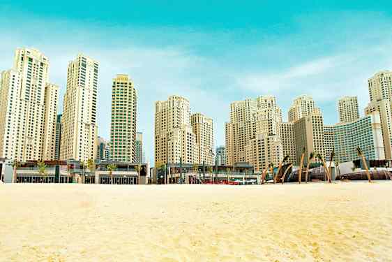 En strand med sand i förgrunden och en rad höga moderna byggnader i bakgrunden. (AI-genererad text)