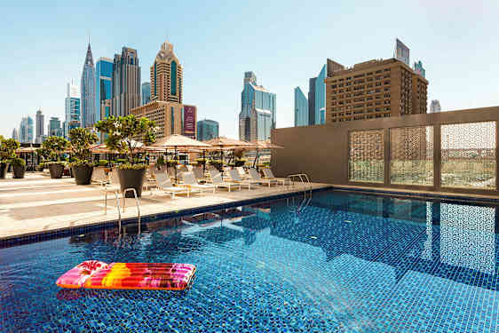 En utomhuspool med uppblåsbar badmadrass, omgiven av solstolar och parasoll. Höga byggnader syns i bakgrunden. (AI-genererad text)