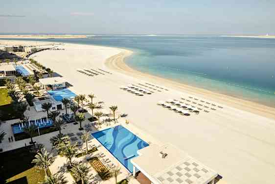 Bilden visar en strand med parasoller och solstolar i raka rader, bredvid en pool och resortområdet omgivna av palmer. (AI-genererad text)