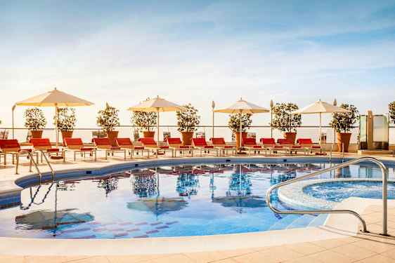 En utomhuspool omgiven av röda solstolar, vita parasoller och krukväxter. (AI-genererad text)