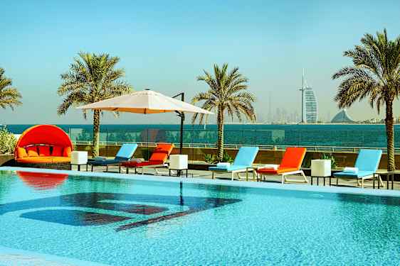 En pool med solstolar och parasoller, omgivna av palmer med havsutsikt och en stadssilhuett i bakgrunden. (AI-genererad text)