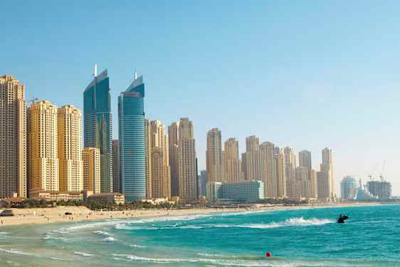 Bild av höghus vid en strand med klart blått vatten, en person är på en jetski nära stranden. (AI-genererad text)