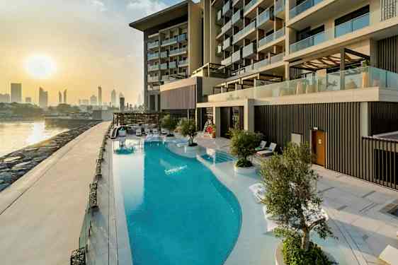 Bild på en lyxig hotellpool vid en byggnad med moderna balkonger. I bakgrunden syns en stadssilhuett och solnedgång. (AI-genererad text)