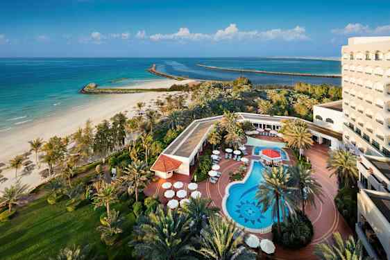 En bild av ett hotell vid stranden med en utomhuspool, solstolar och omgivande grönska. (AI-genererad text)